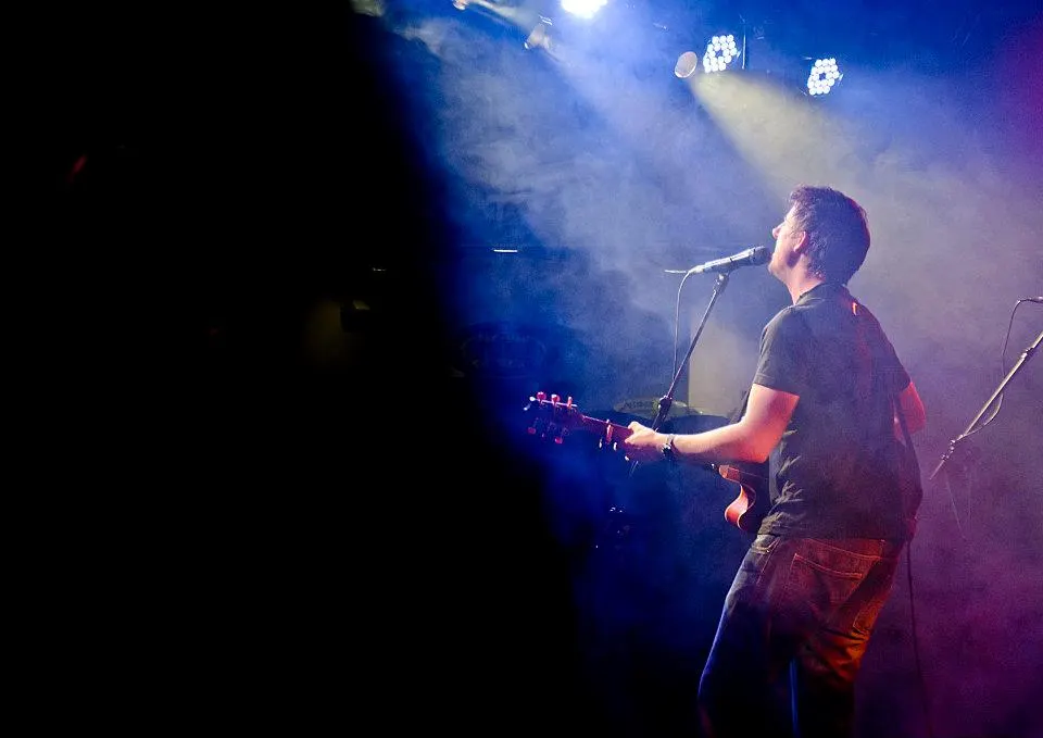 Ryan Inglis on a stage lit in blue, singing with his guitar
